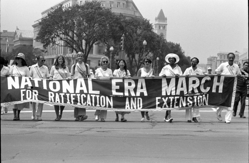 Their banner reads 'National ERA March for Ratification and Extension. Visible at center is the curved facade of the Federal Trade Commission building and, behind it, the tower of the Old Post Office Pavilion.