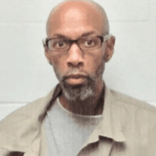 Image description: a mugshot of a Black man with a beard wearing glasses and a khaki shirt