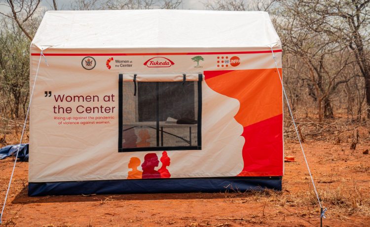 A tent stands on a red dirt ground, beneath trees. It has “Women at the Centre” written on the front, with the logos of UNFPA, Takeda and others